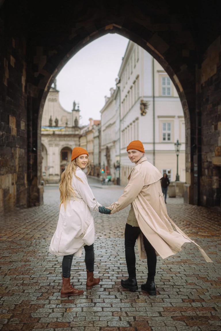 Mladý pár v béžových kabátoch sa drží za ruky pozerajúc za seba na Karlovom moste v Prahe.